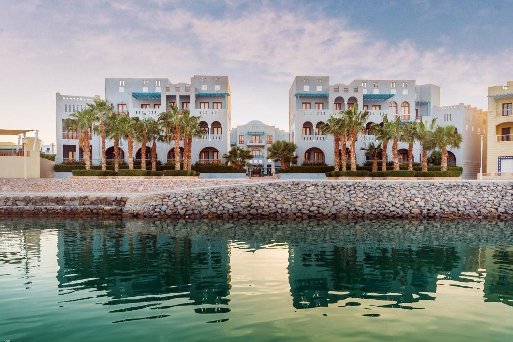 Coastal resort buildings with palm trees along a stone seawall, reflections shimmering in the calm water.