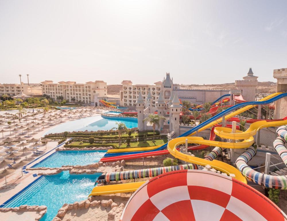 Wide view of a resort water park with colorful twisting slides, castle towers, and multiple pools surrounded by sun loungers and umbrellas, under a bright sky.