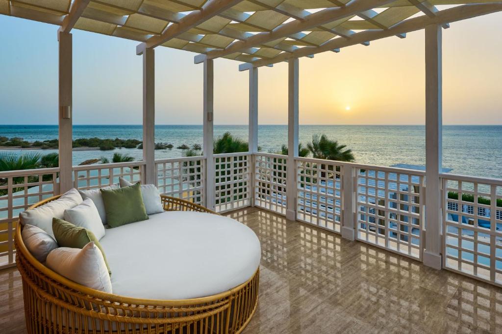 Round wicker daybed with white cushions and green pillows on a balcony overlooking the ocean at sunset.