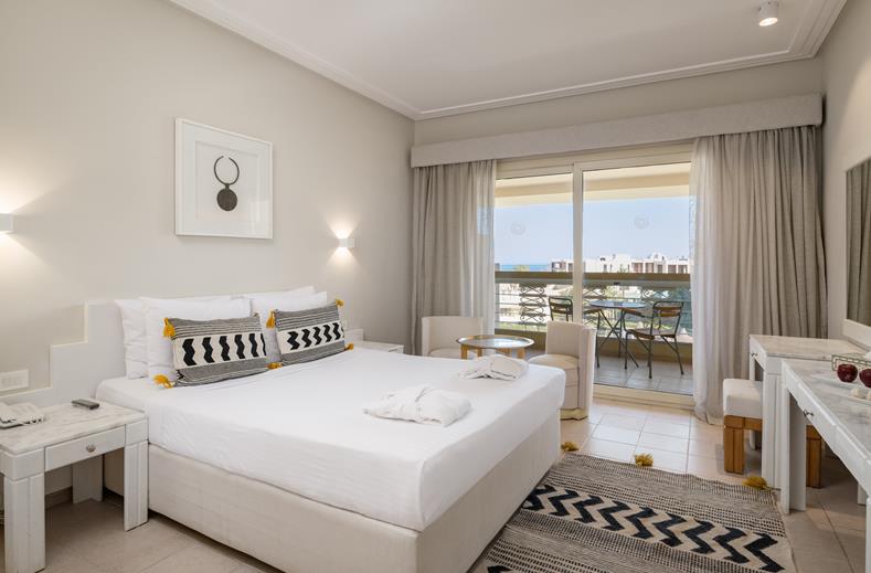 Hotel room with a large white bed, nightstands, and a balcony overlooking the sea.