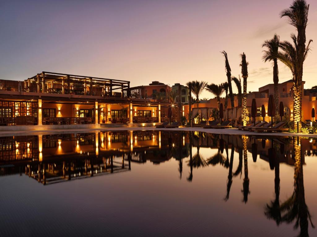 Sunset-lit hotel resort with an illuminated terrace and palm trees around a calm pool, reflecting the building and trees in the water.