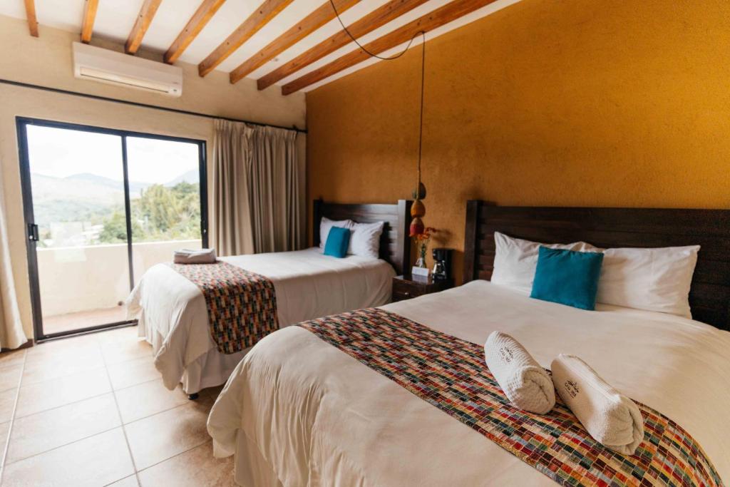 Hotel room with two double beds, teal pillows, and colorful runners; balcony sliding door to mountain view on a warm-toned wall.
