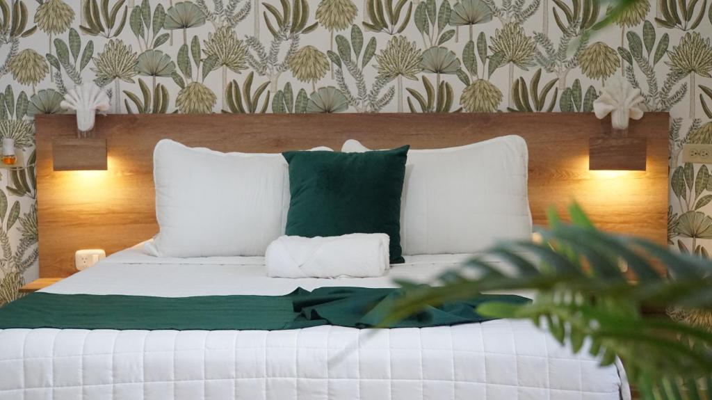 A neatly made bed with white bedding, a dark green throw and matching pillow atop a wooden headboard, flanked by wall lamps and botanical wallpaper.