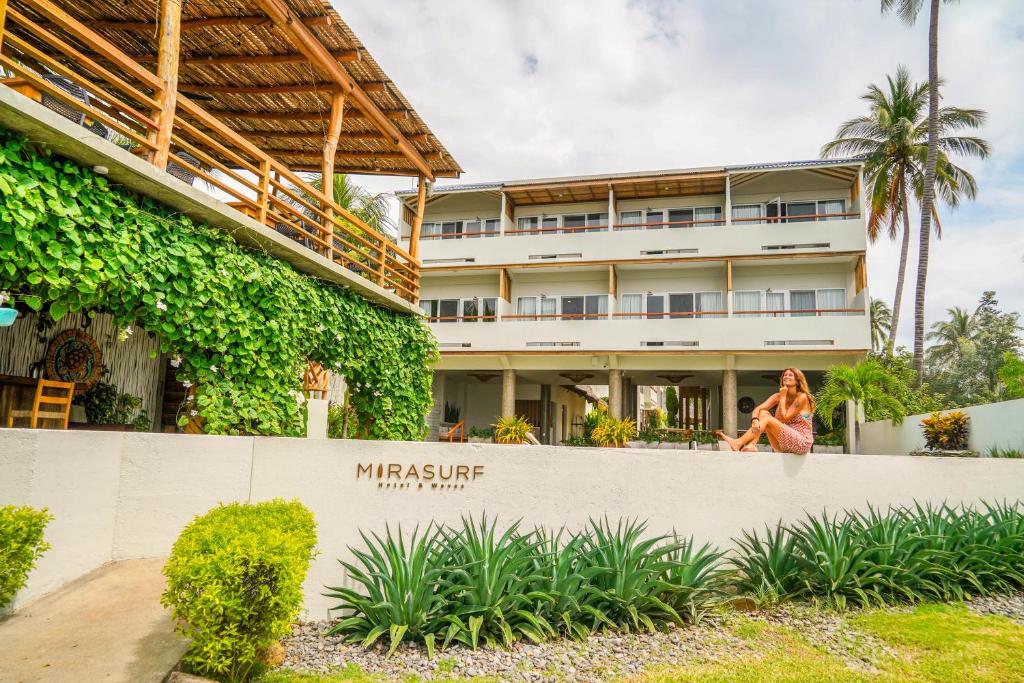 Front of a tropical hotel with white balconies, lush greenery, and a woman sitting on a low wall in front near the MIRASURF sign.