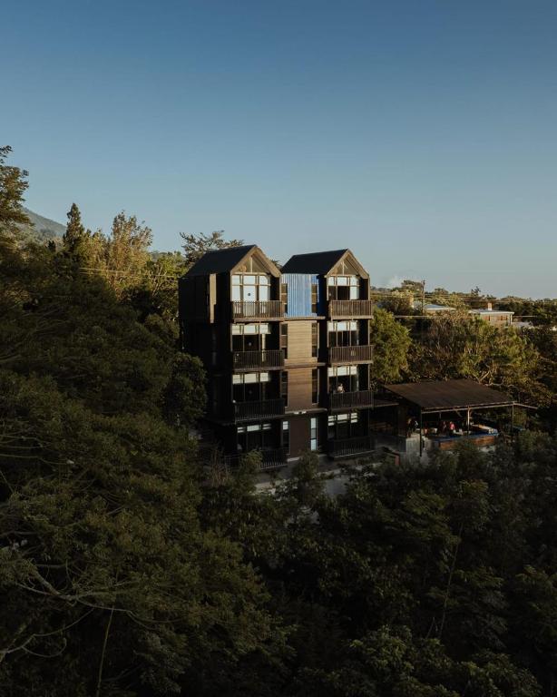 Wood-clad apartment building nestled among trees on a hillside under a clear blue sky, with a shaded outdoor area to the right.