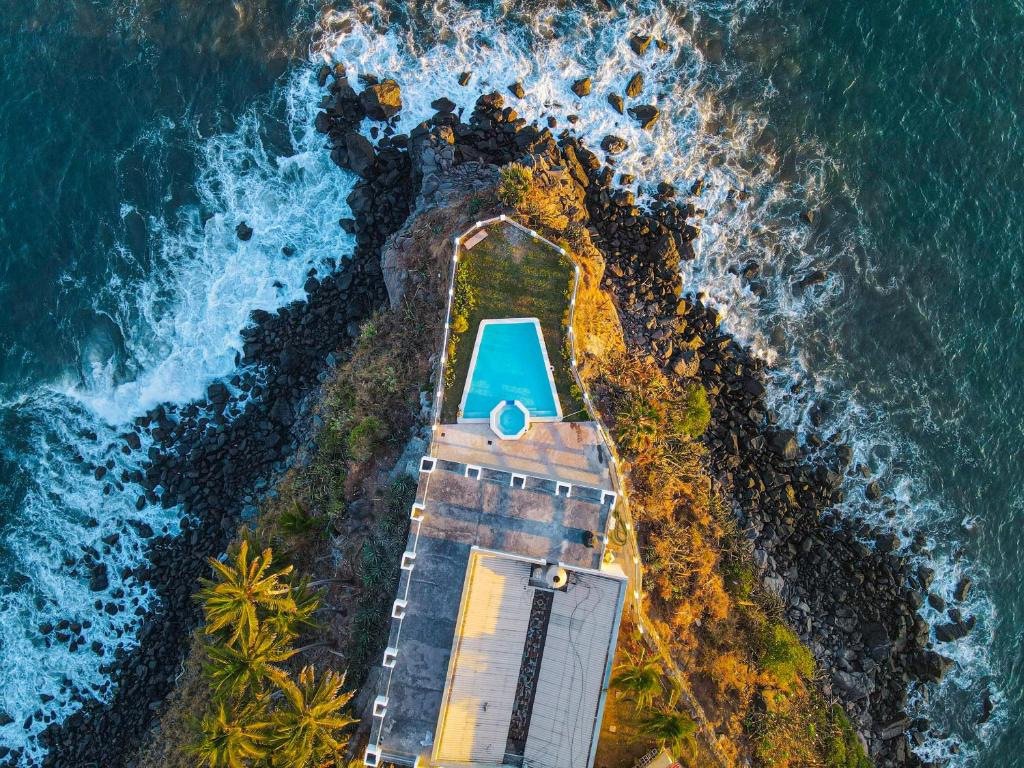 Aerial view of a turquoise pool on a rocky promontory jutting into the sea, with waves crashing around the rocks and palm trees nearby
