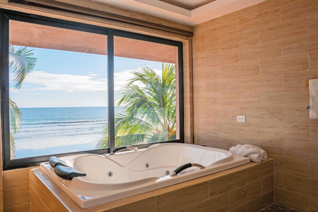 White jetted tub in a tiled bathroom, with a large window showing a sandy beach and palm trees outside.