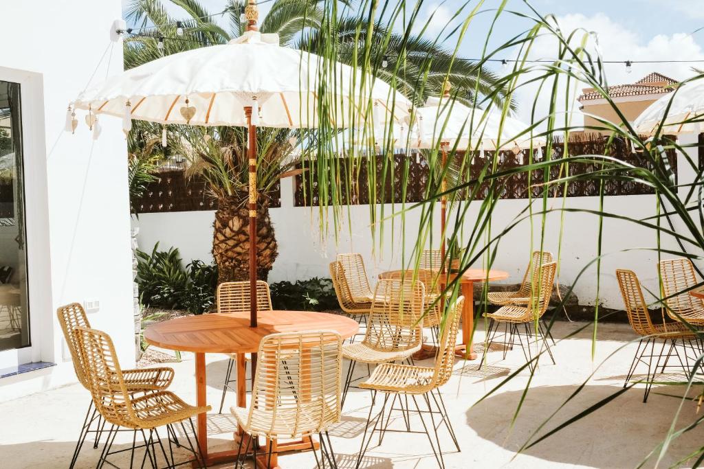 Outdoor patio with round wooden table, beige wicker chairs, and white umbrellas in a sunny courtyard.