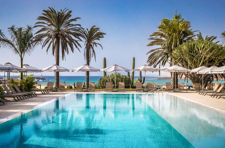 Resort pool area with turquoise water, lounge chairs, and white umbrellas under palm trees by the ocean.