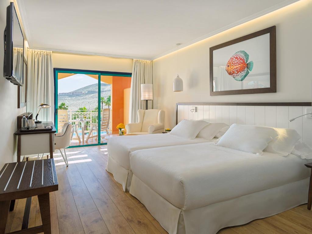 Bright hotel room with two white beds, a wall-mounted TV, and a balcony view through glass doors to orange exterior seating area.