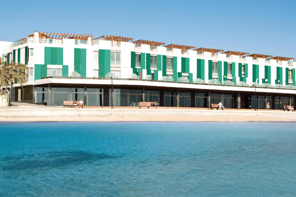 Modern white hotel with green vertical facade panels along a beachfront promenade; clear blue water in the foreground and a few benches nearby.