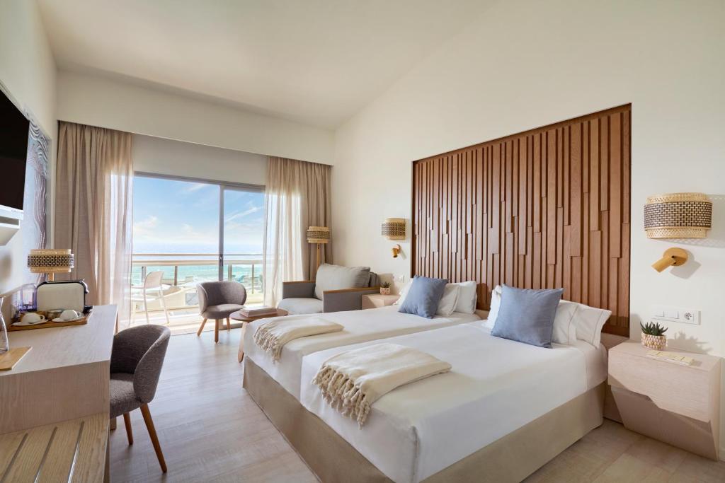 Hotel room with two sea-view beds, cozy seating area, and balcony overlooking the ocean.