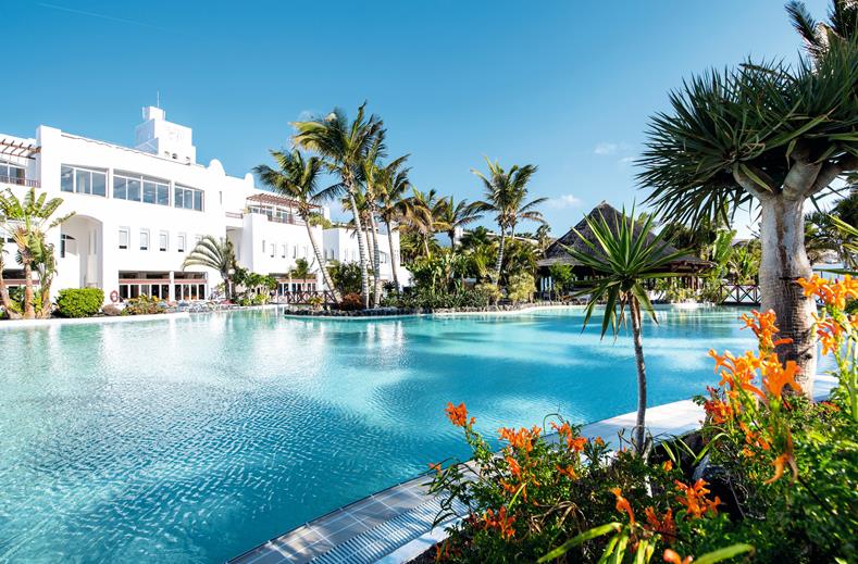 Resort pool area with clear blue water, white buildings, and palm trees under a bright sky.