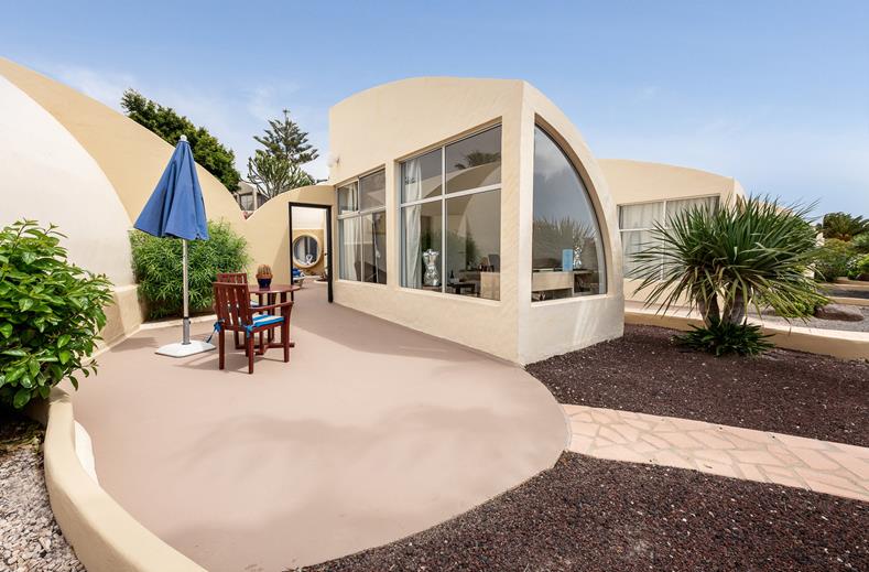Curved beige house with large arched windows, patio seating under a blue umbrella on a sunny day