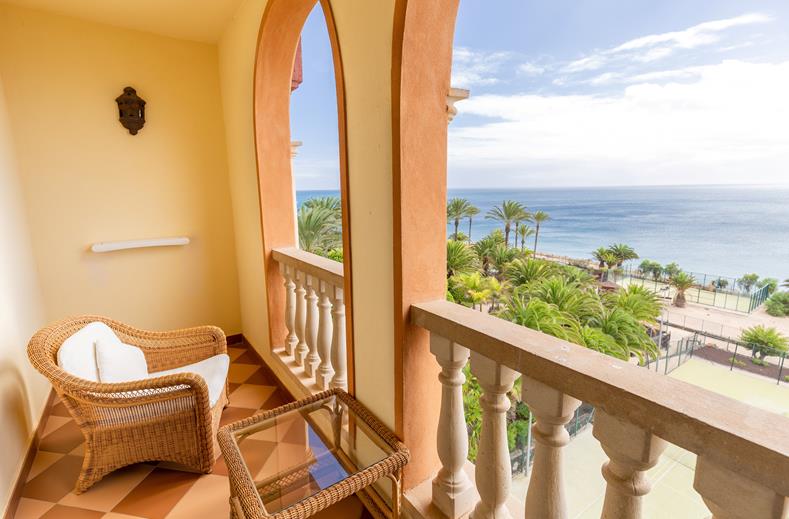 Wicker chair and glass-topped table on a tiled balcony, with arched openings and a view of the ocean and palm trees beyond.