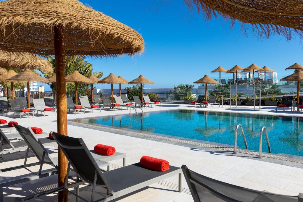 Outdoor resort pool area with blue water, straw umbrellas, and gray lounge chairs with red towels.