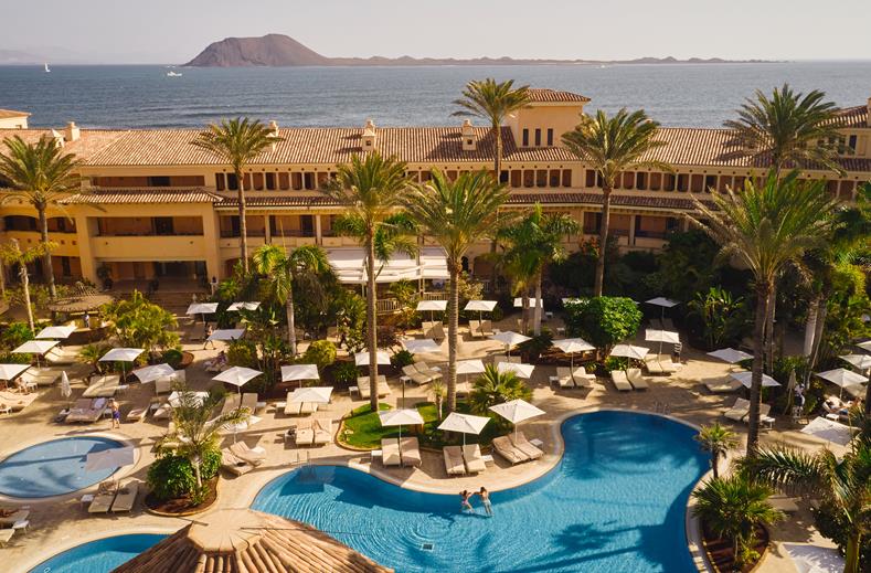 Aerial view of a resort pool area with lounge chairs, palm trees, and a large building, ocean and island in the background.