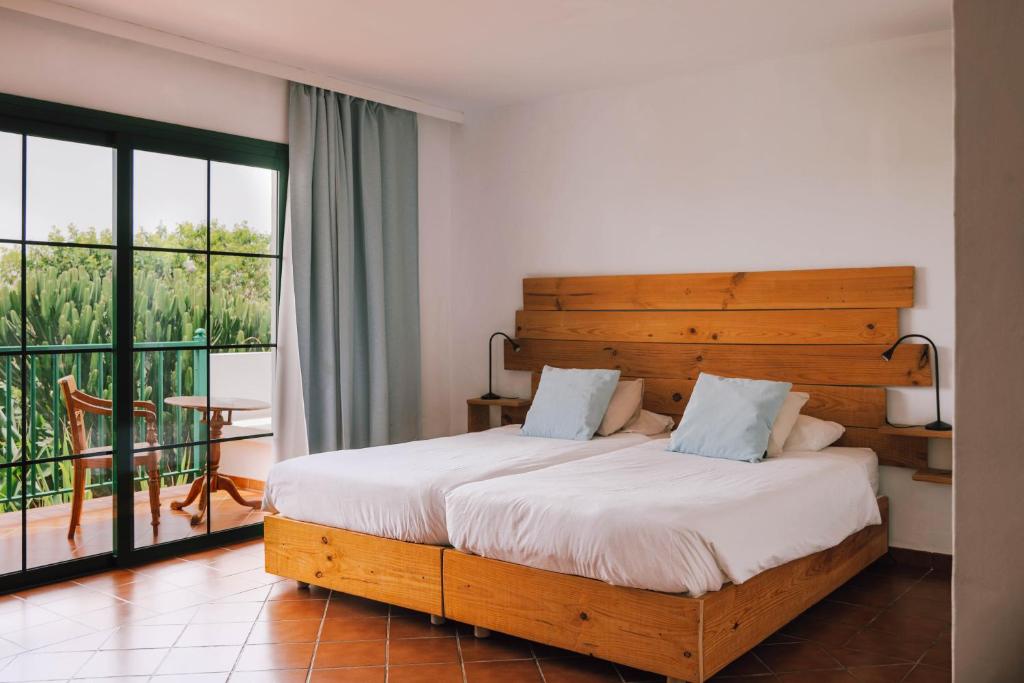 Two twin beds with white linens and light blue pillows sit side by side against a wooden headboard in a sunlit hotel room; a balcony with glass doors and outdoor chairs is visible beyond, and terracotta tile floors complete the scene.