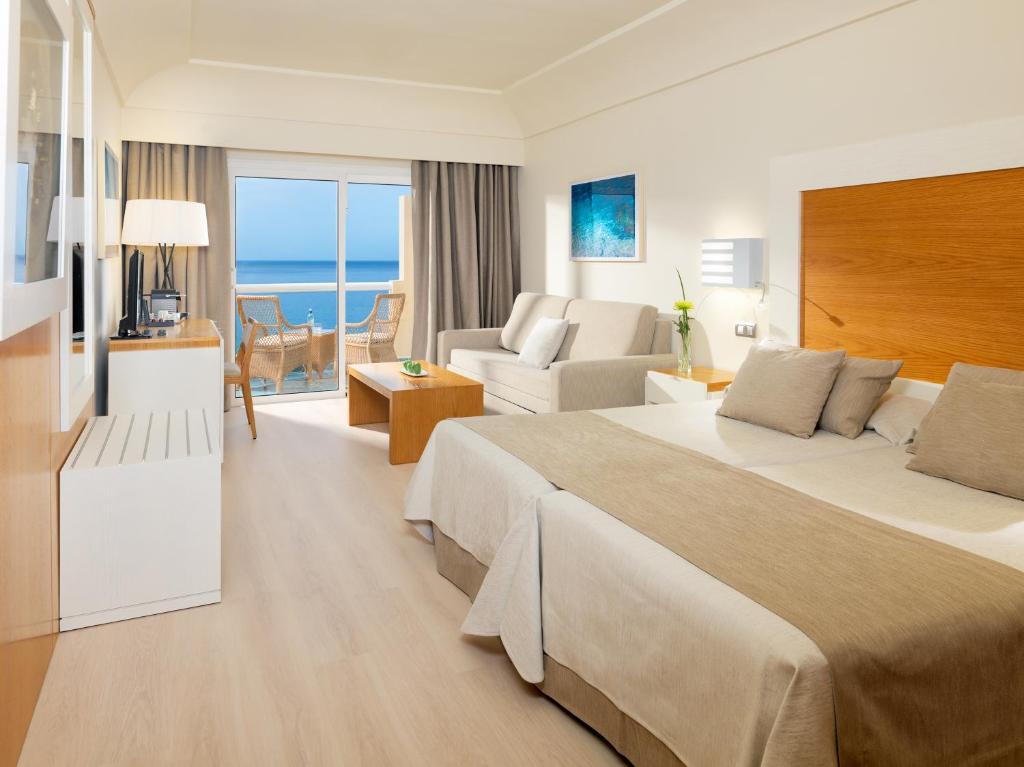 Hotel suite with two large beds, neutral décor, and a balcony view of the sea; seating area and a coffee table in front of a sofa.