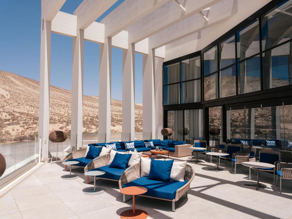 Open-air lounge terrace with white pillars, blue cushioned seating, and small round tables overlooking desert hills and clear blue sky near a glass-walled building.