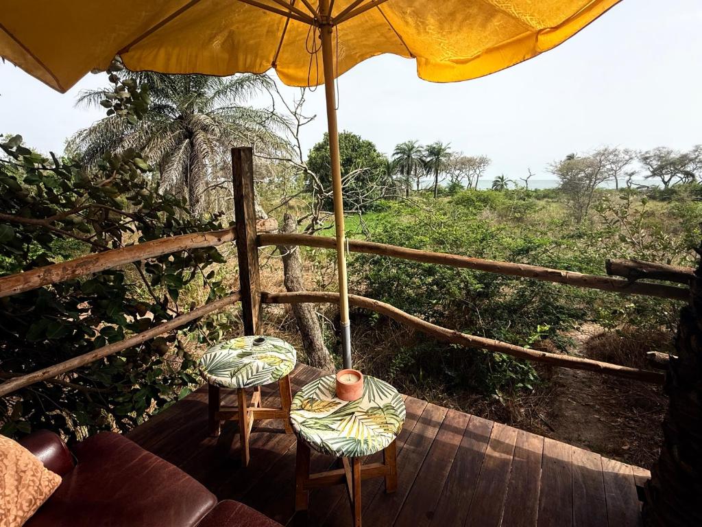 Outdoor wooden deck with a yellow umbrella, two small tropical-pattern tables, and stools overlooking a leafy tropical landscape.