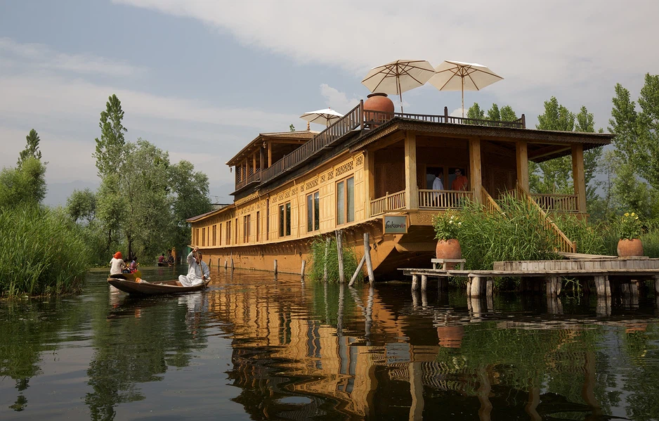 Large wooden houseboat on a calm river with a two-level deck, railings, and white umbrellas; passengers on board with trees in the background.
