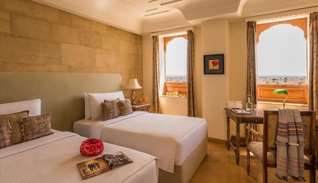Twin‑bed hotel room with beige decor, nightstands, and a desk by arched windows framing a desert view.