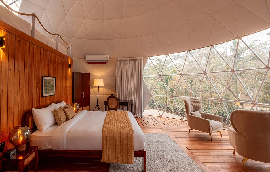 Geodesic-dome hotel room with a king bed, wooden headboard, and a seating area by large glass walls overlooking trees outside.
