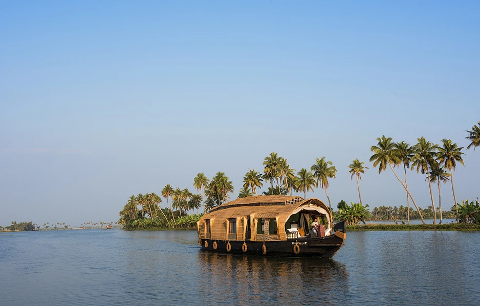 Traditional thatched-roof houseboat gliding on a calm backwater, with a palm-lined riverbank in the background.