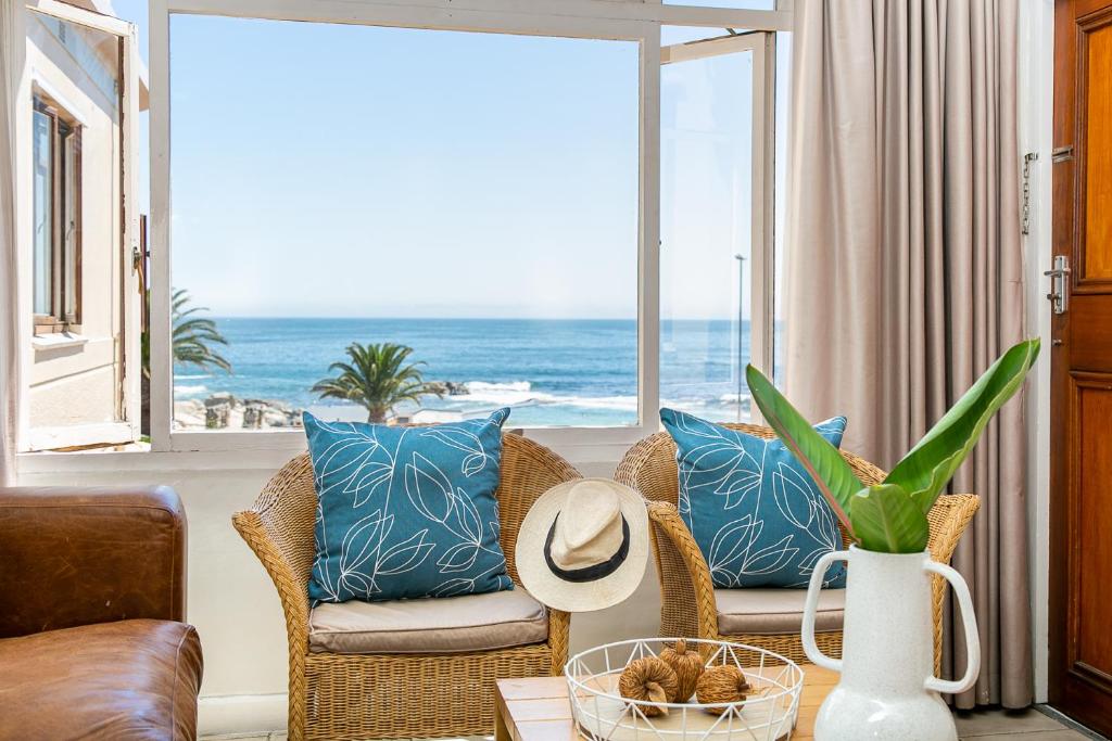 Sunlit seaside living area with two wicker chairs and blue leaf-pattern cushions facing the ocean through a large window; a hat rests on one chair and a white pitcher with a green plant sits on the table.