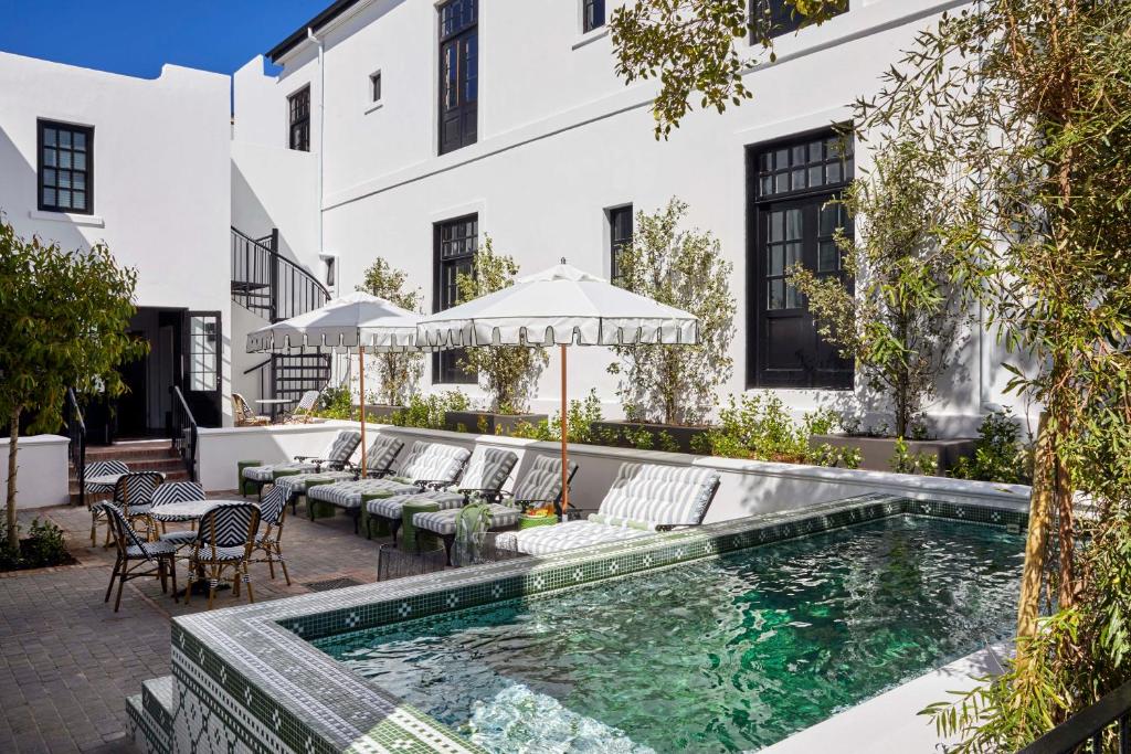 White-walled hotel courtyard with a rectangular pool, striped lounge chairs, and white umbrellas along the pool edge.