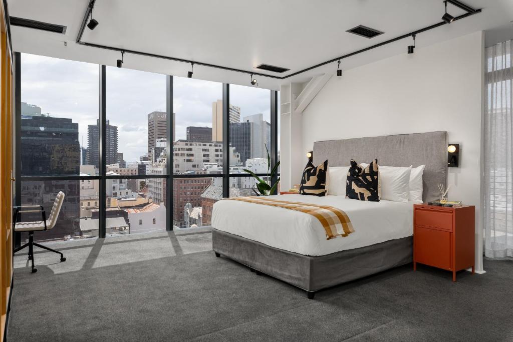 Modern hotel bedroom with a large windowed wall overlooking the city, a grey upholstered bed, and an orange nightstand on the right side.