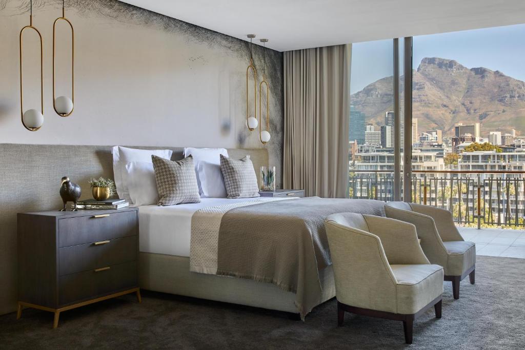 Spacious hotel room with a king bed, two nightstands, and a seating area, overlooking a city and mountain landscape from a balcony window.
