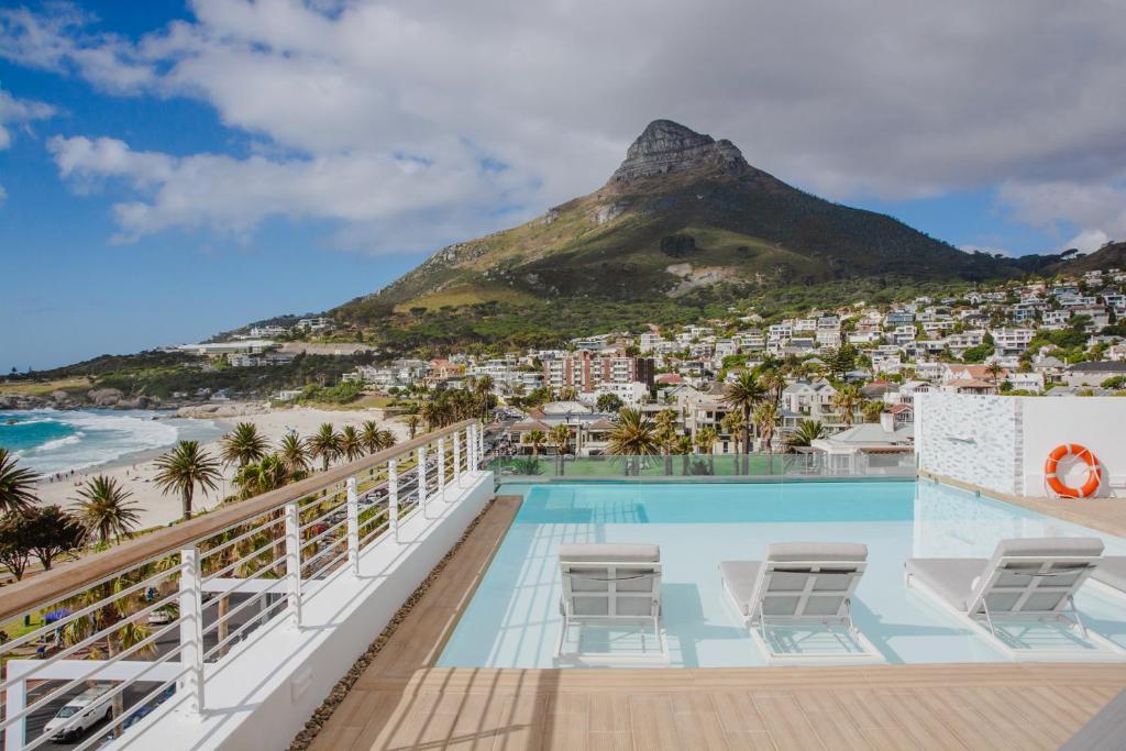 Rooftop pool with white lounge chairs overlooking a seaside town, palm trees, and a tall mountain in the distance.