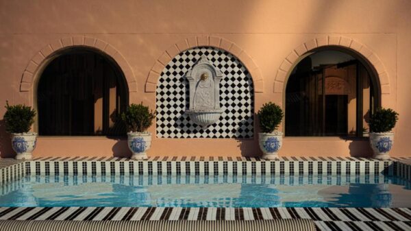 Terracotta wall with three arches, a central decorative fountain on a black-and-white tiled backdrop, and potted shrubs flanking a blue-tiled pool area.