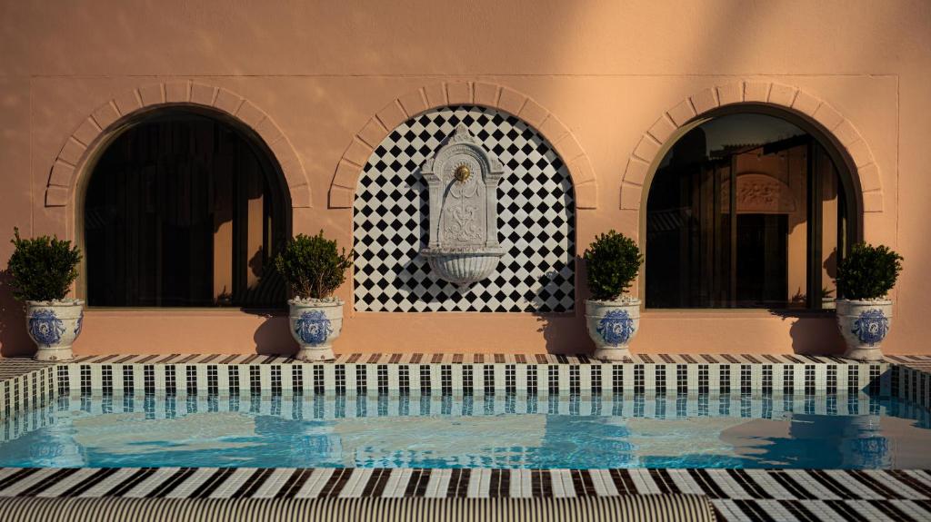 Terracotta wall with three arches, a central decorative fountain on a black-and-white tiled backdrop, and potted shrubs flanking a blue-tiled pool area.