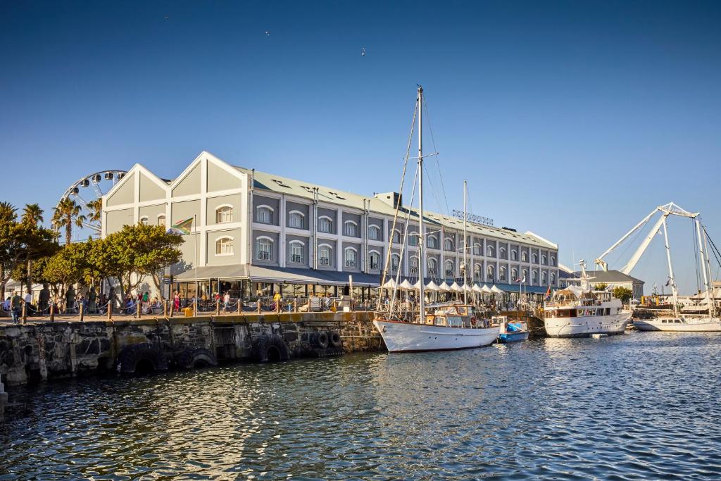 Harborfront with a long gray, arched-window building beside the quay; sailboats docked along the water under a clear blue sky.