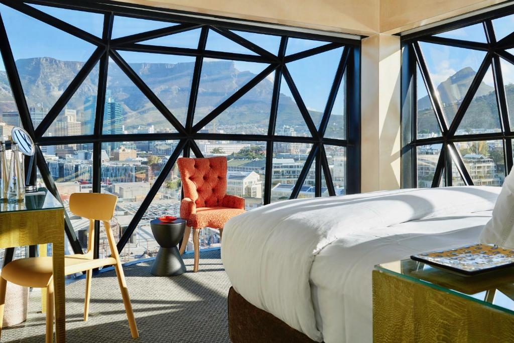 Hotel room with a large geometric glass wall showing a city and mountain view, a white bed, and a yellow chair with an orange armchair nearby.