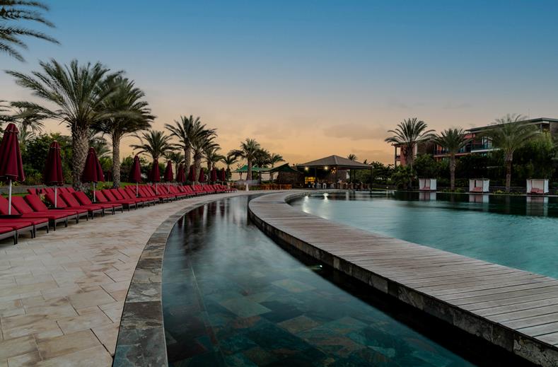 Resort pool with curved wooden walkway, red lounge chairs, and palm trees at sunset