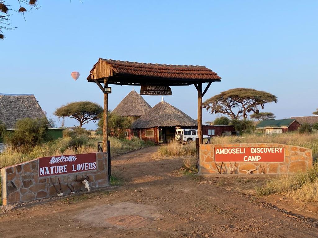 Entrance to Amboseli Discovery Camp with welcome signs and thatched huts in a savannah setting.