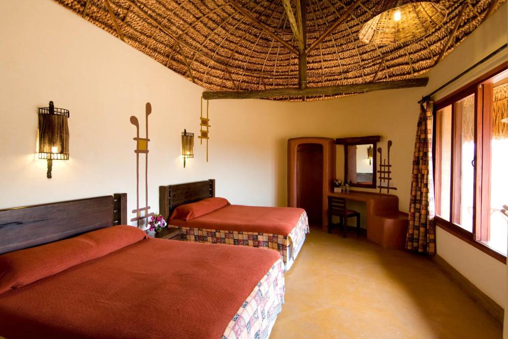 Two twin beds with red bedding in a rustic thatched-roof hotel room, warm lighting, and wooden furniture near a large window.