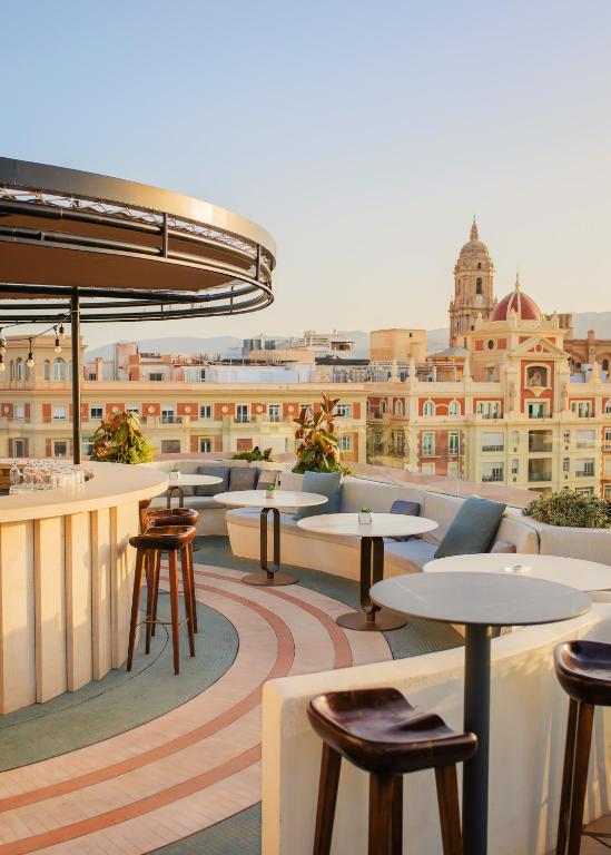 Rooftop bar and seating area with round white tables and wooden stools, overlooking a sunlit cityscape with domed church towers in the distance.