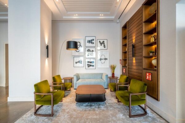Contemporary hotel lounge with four olive-green armchairs around a wooden coffee table, and a light blue sofa against a wall of framed abstract art.