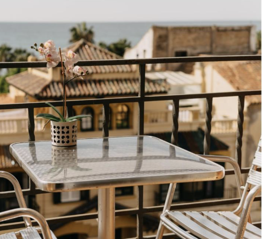 Metal patio table and chairs on a balcony overlooking rooftops and the sea, with a potted pink orchid.