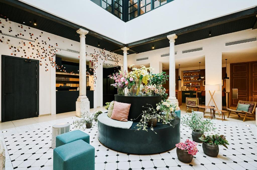 Hotel lobby with a circular black sofa centerpiece filled with flowers and cushions, surrounded by plants and white tiled floor.