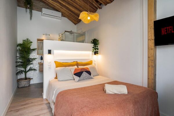 Cozy modern bedroom with white walls, a beige bedspread, orange and gray pillows, and a folded towel on the bed; a large potted plant sits in the corner, with a pendant light and wooden ceiling beams overhead, and a Netflix screen on the wall to the right.