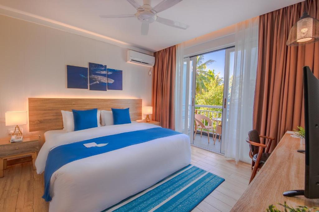 Hotel bedroom with a king bed, white linens, blue pillows and throw, and a balcony with white chairs seen through glass doors outside the room central to a tropical view.