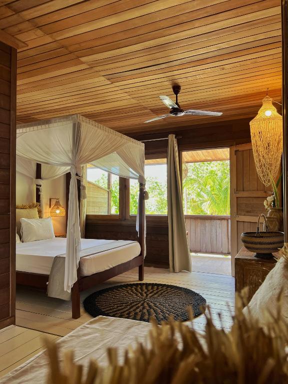 Tropical bedroom with a four-poster bed draped in white fabric, wooden walls, a ceiling fan, and an open veranda view of greenery.