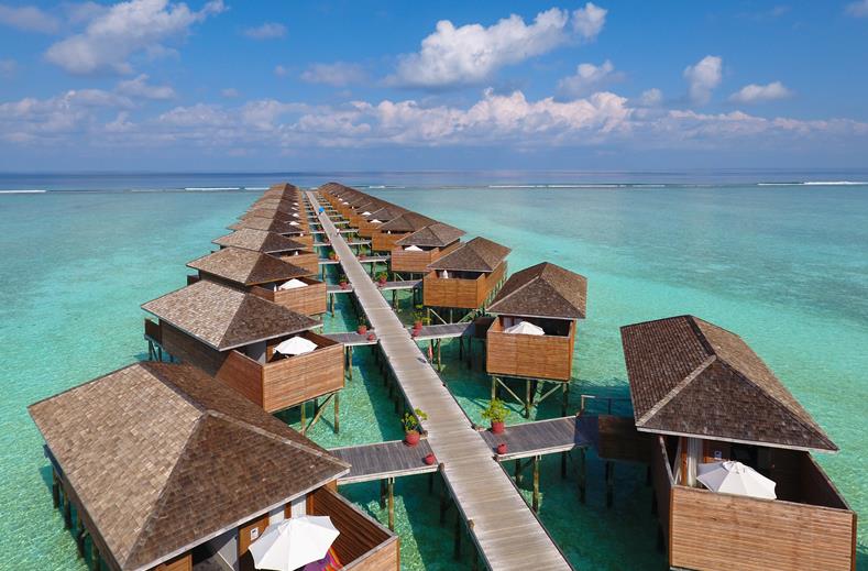 Overwater bungalows connected by a long wooden boardwalk above turquoise sea under a blue sky.