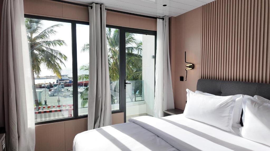 Hotel room with a large window view of palm trees and the beach; sunlit bed with white linens and a grey headboard.
